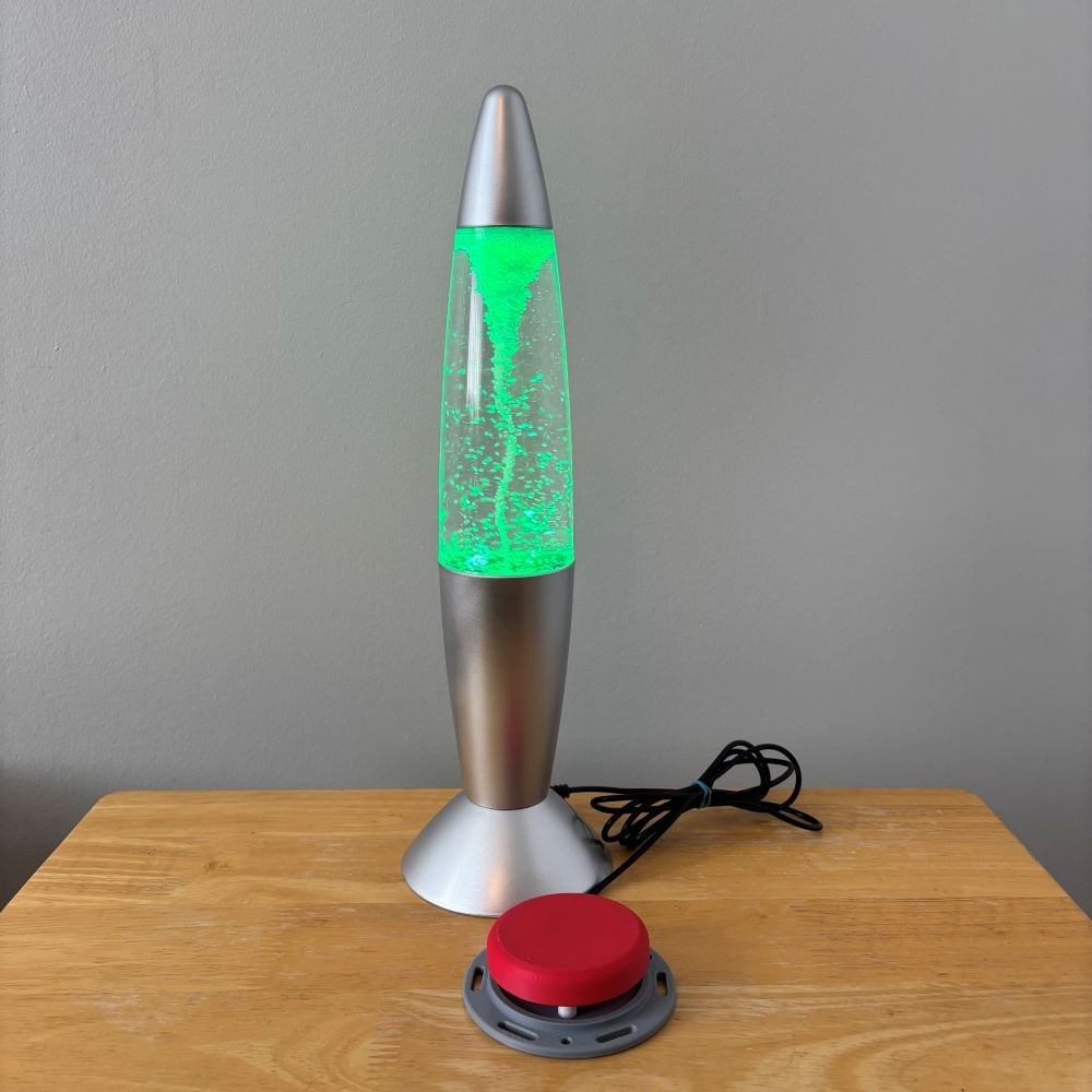Switch adapted silver lamp toy that is lit up green and has a tornado effect inside. The lamp looks like a lava lamp but is a tornado inside instead. There is a red assistive switch plugged into the lamp.