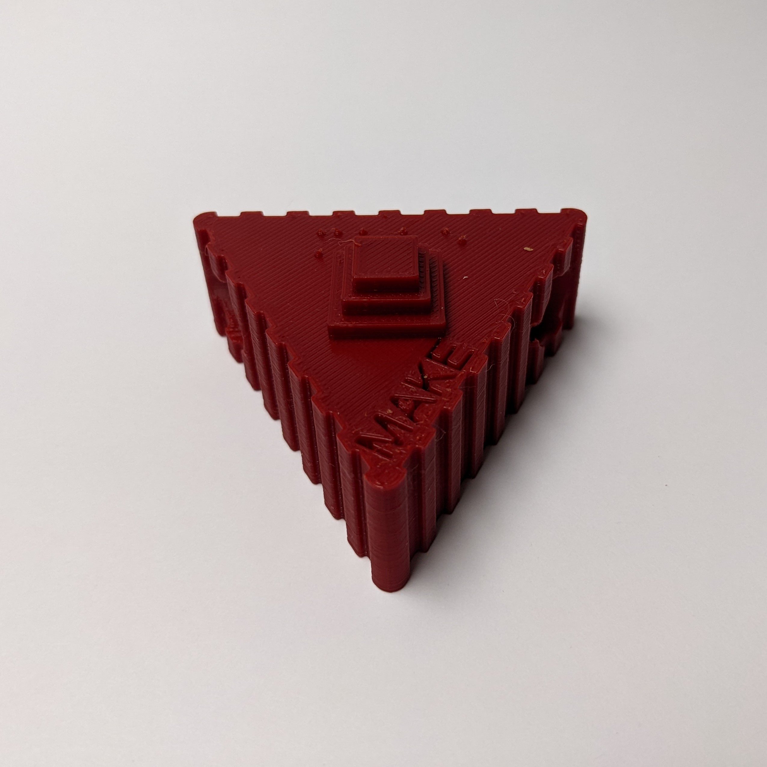 A red 3d printed project core symbol of the 'Make' symbol against a white surface.. The symbol consists of a red triangle shaped base with a vertically ridged texture on the side. On the top of the symbol, the word 'Make' is engraved, 'Make' is written in Braille, and there is a symbol of a three-tiered box.