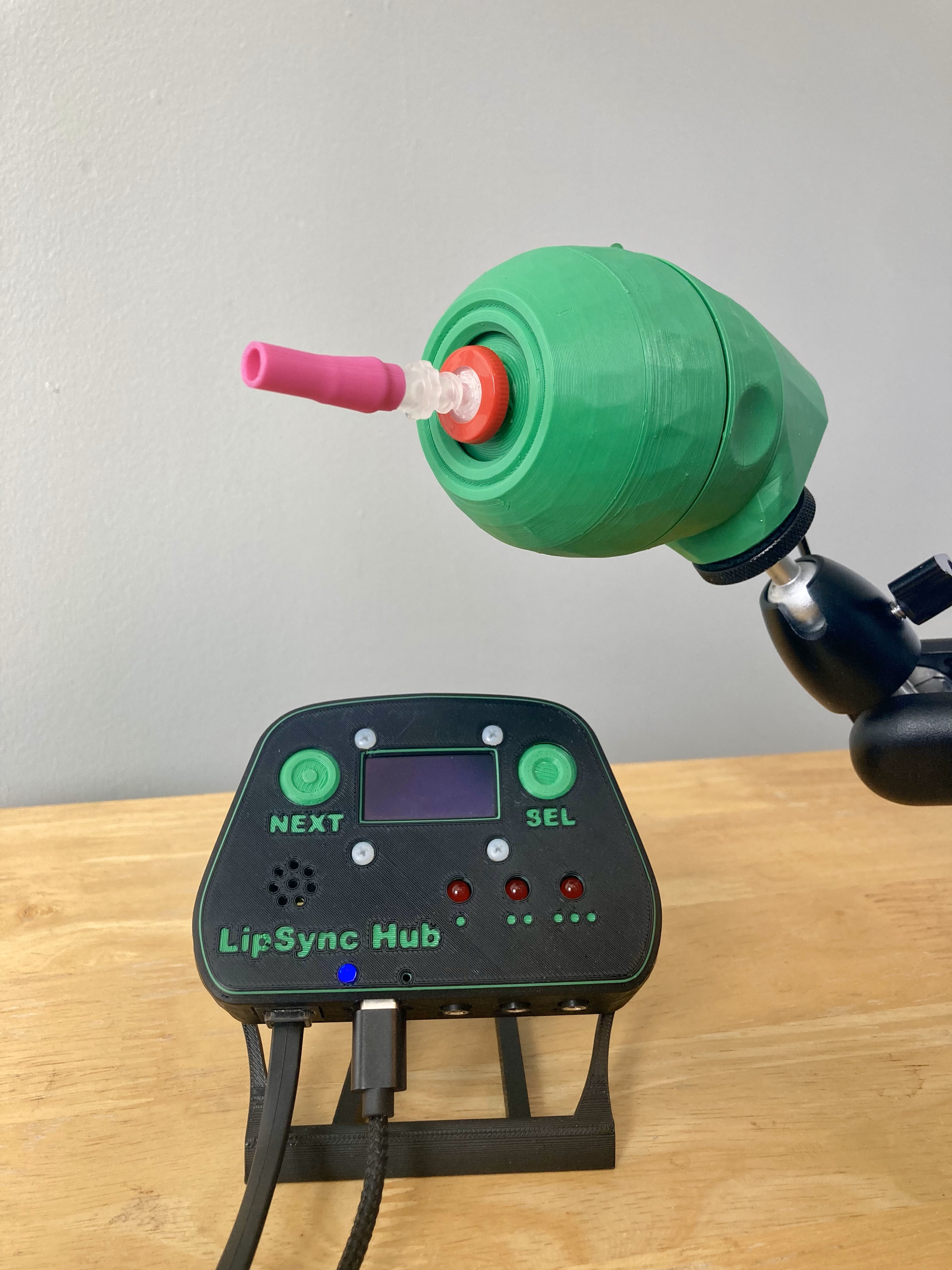 A green 3D printed LipSync joystick with a pink mouthpiece, mounted and shown above a black LipSync Hub on a table.