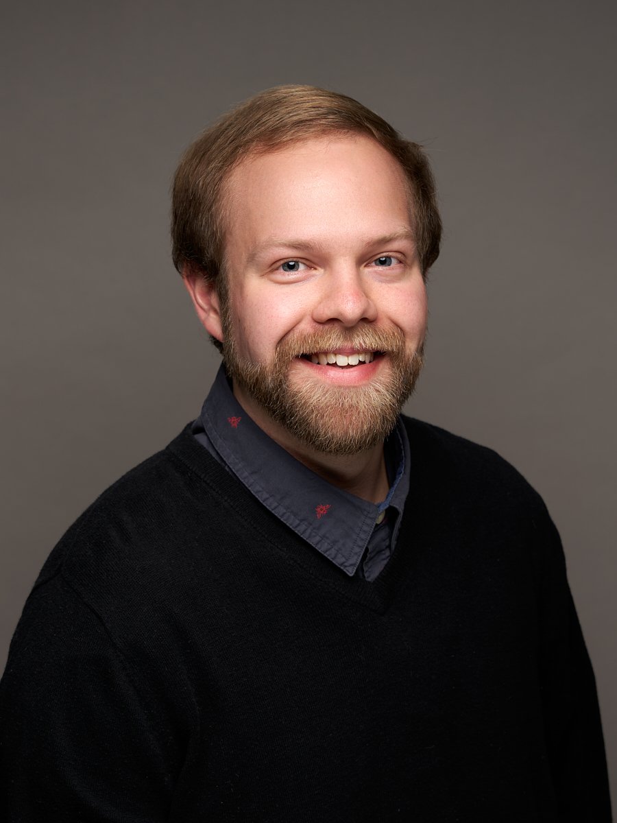 A man with blonde hair and mustache wearing a blue sweater against a grey background.
