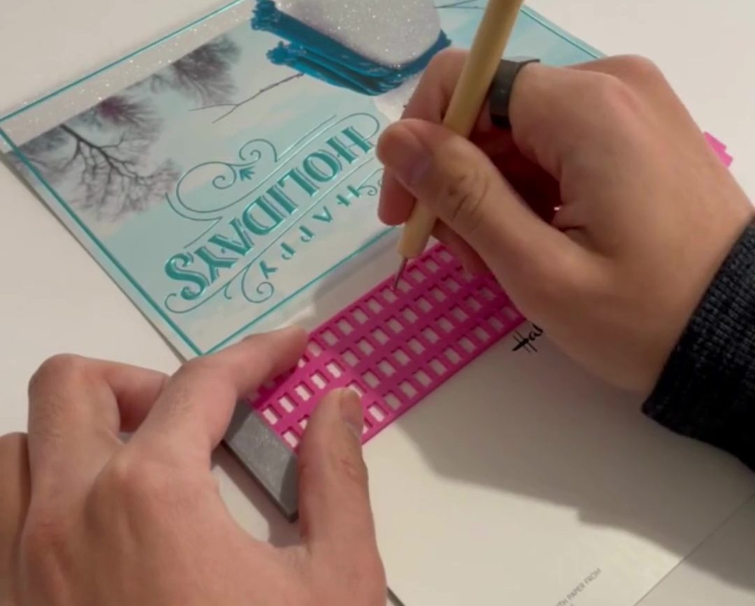 A pair of hands embossing braille on a holiday card using the 3d printed braille and stylus set