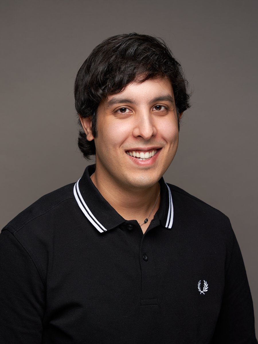 A man with wavy black hair and black polo shirt against a grey background.