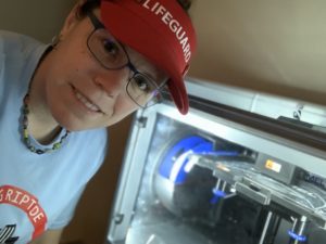A volunteer with a red hat on that reads Lifeguard taking a picture beside a 3D printer.