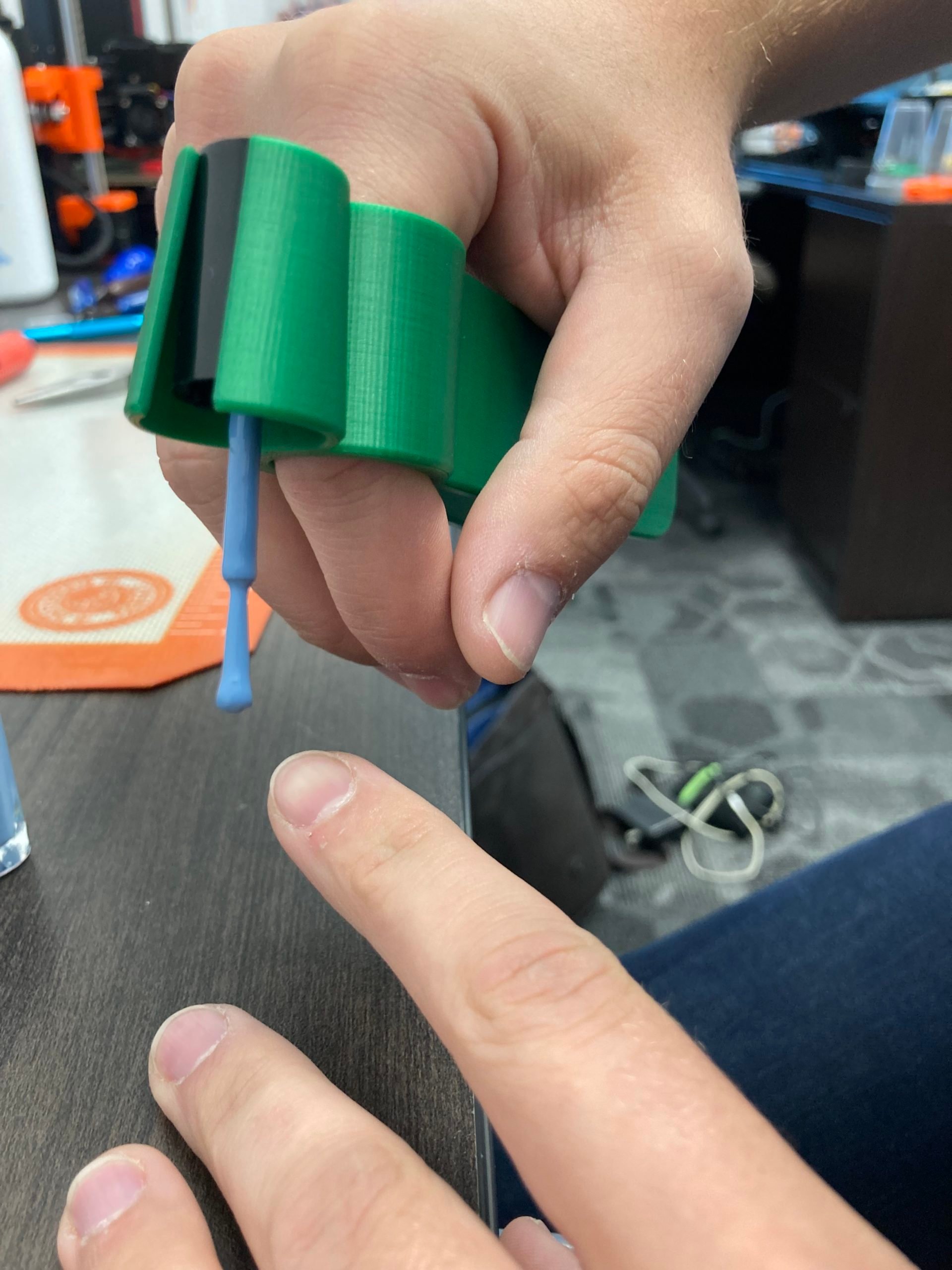 A finger looped through a green 3d printed assistive nail polish applicator with a blue brush attached. Underneath the brush is finger ready for polish application.