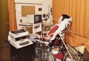 Man in a wheelchair with a straw-like device mounted near his mouth, surrounded by several computers