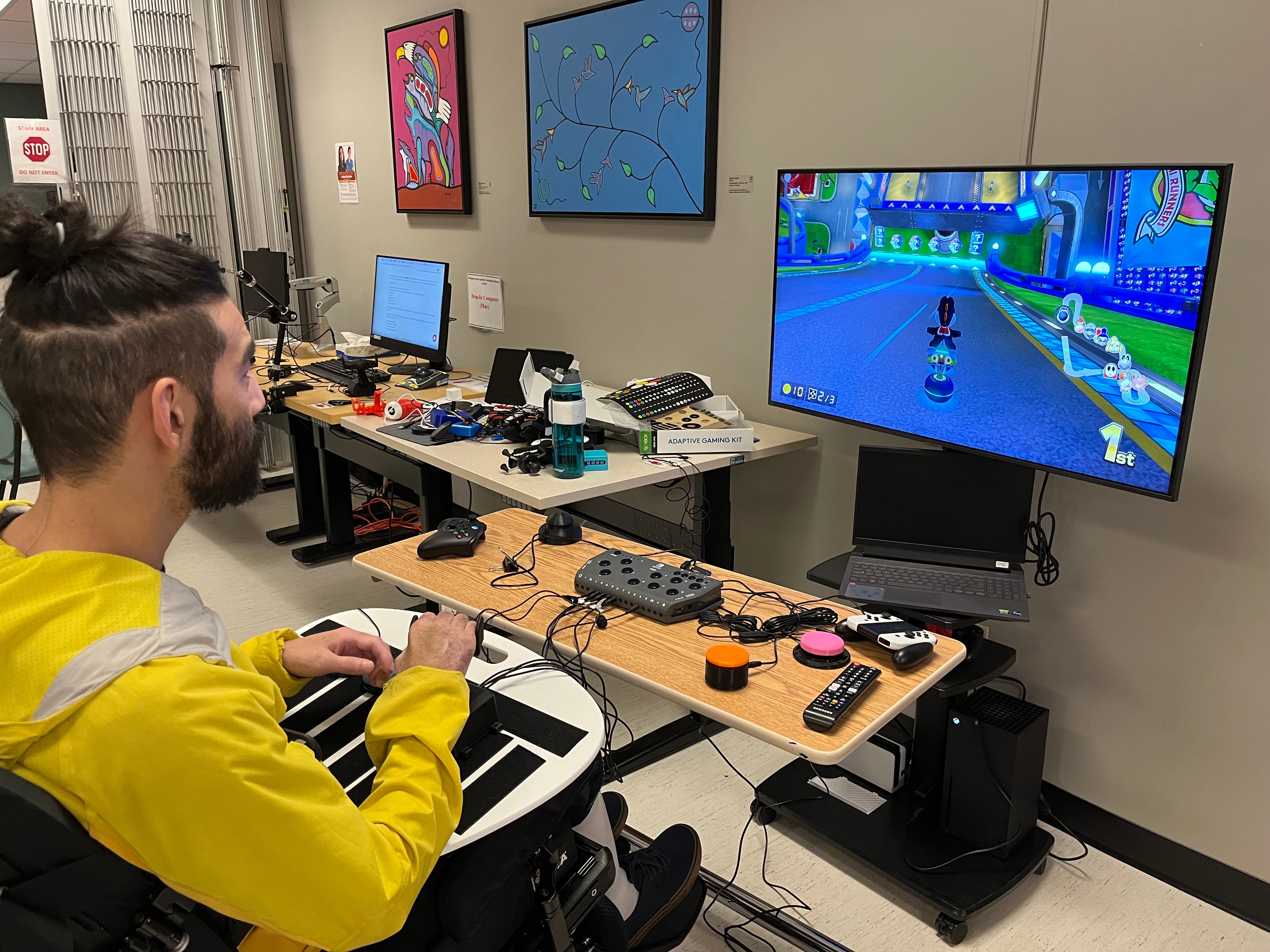 A person in a wheelchair playing a Mario Kart with an adaptive joystick and assistive switch on a hook and loop lap tray. various other assistive technologies are shown on the table.
