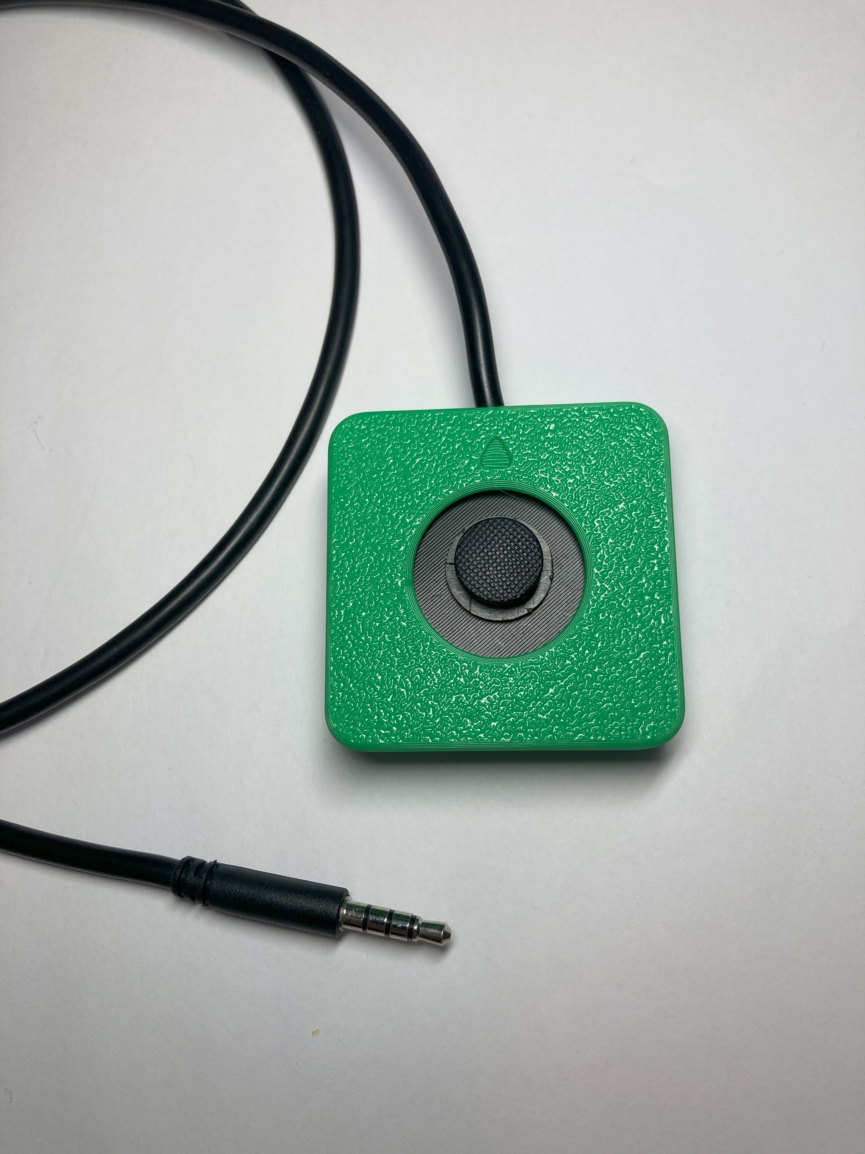 Green Aspen Sliding Joystick, from above. The joystick enclosure is square, green, and 3D printed. In the middle is a black circle and a darker black circle inside of that, which is the joystick topper. A black cable comes out of the back of the joystick and wraps around to show the TRRS jack, which looks like a headphone cable jack.