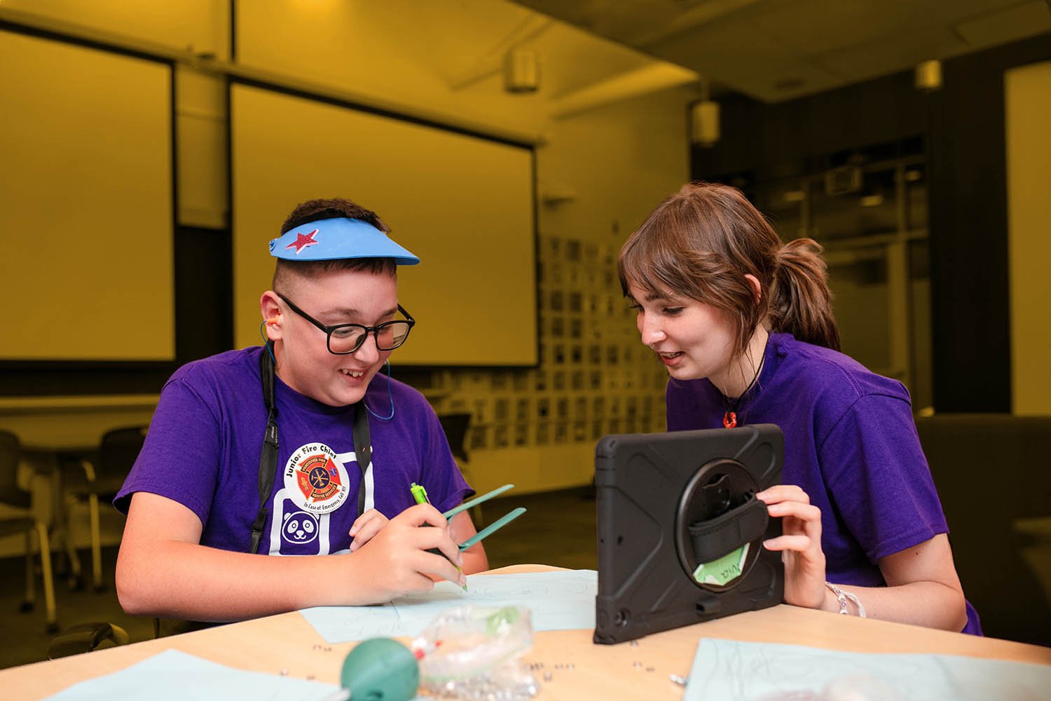 A boy writing with a green writing aid with a woman looking at his writing.