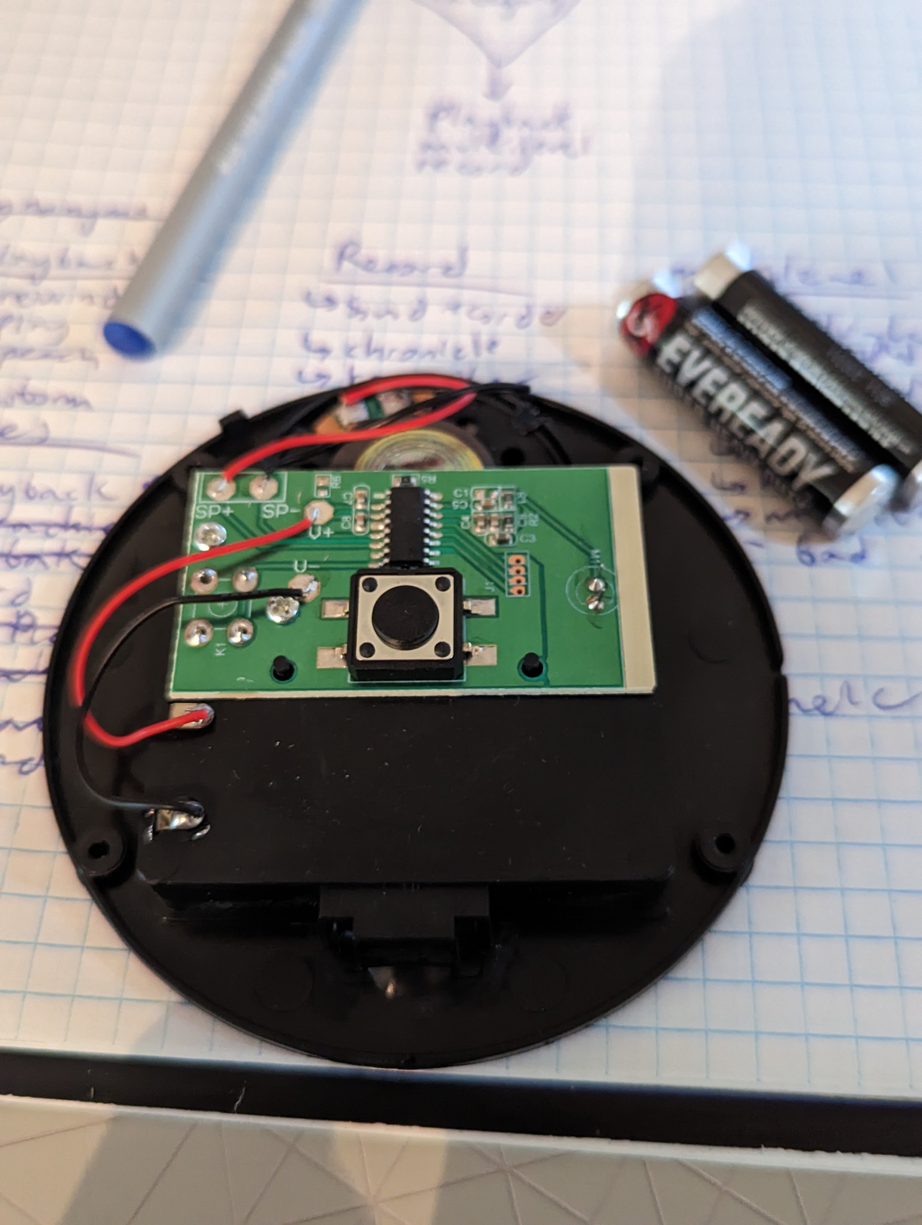 A black, round switch base sitting on top of graphing paper.  There is a green printed circuit board in the center of the black base with red and black wires connected to it.