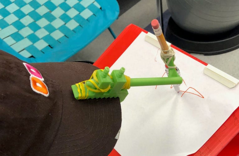 An assistive device attached to a hat to hold a pencil.
