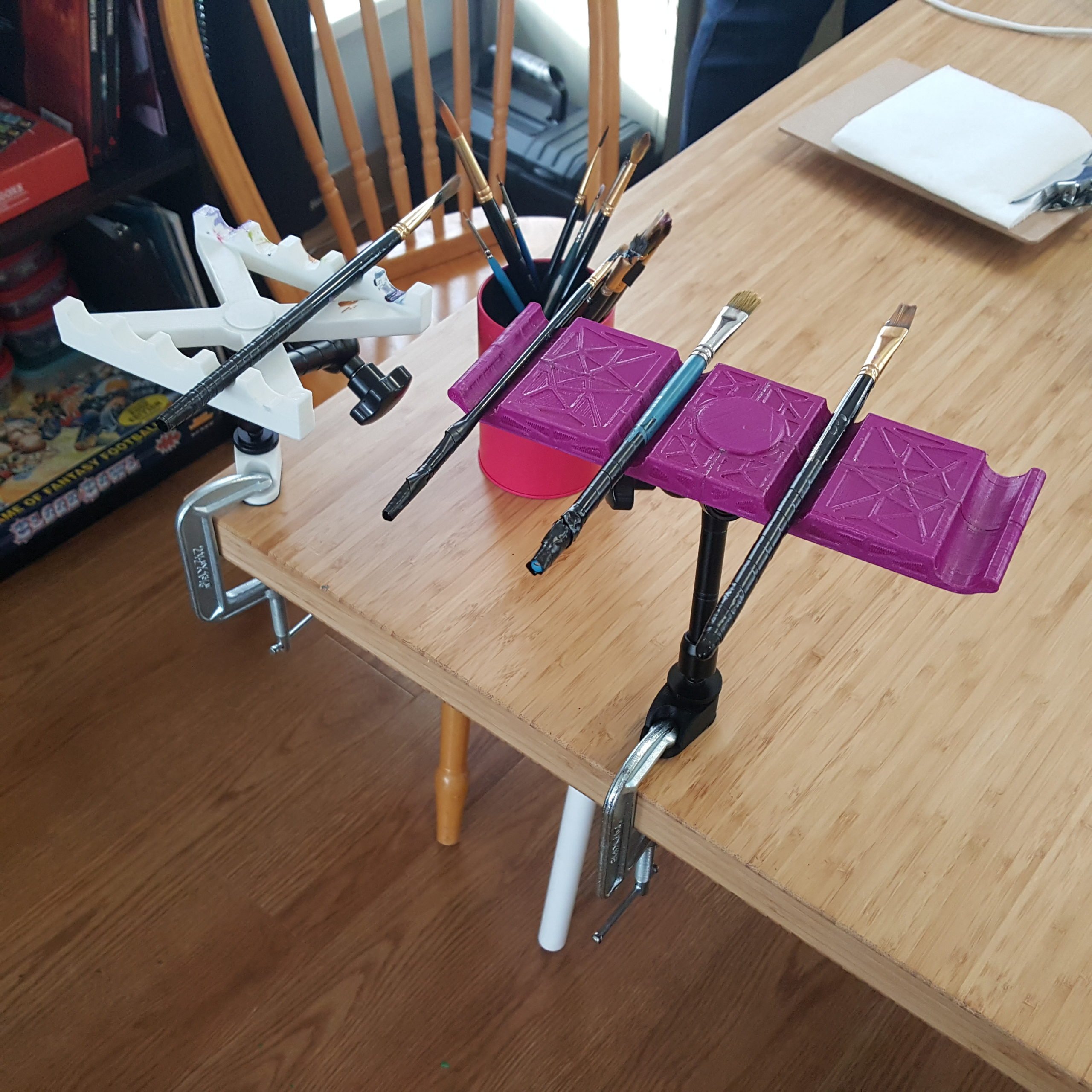 Pink and white 3d printed paintbrush stands holding several paintbrushes. The stands are mounted to a wooden table by a black mounting arm.