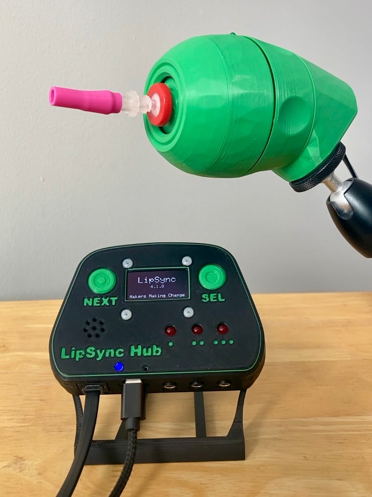 A green 3D printed LipSync joystick with a pink mouthpiece, mounted and shown above a black LipSync Hub on a table. On the Hub screen reads "LipSync v4.1.0 Makers Making Change."