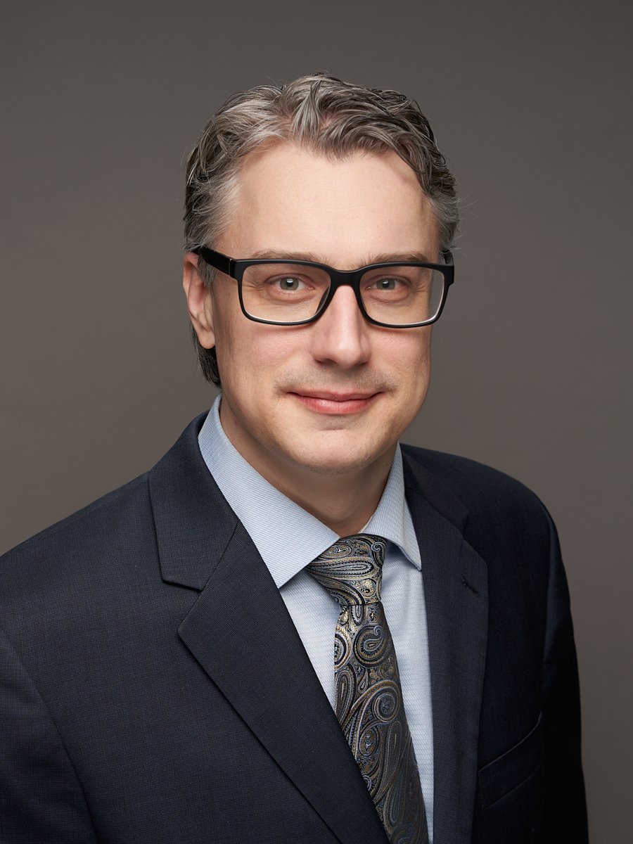 A man with black rimmed glasses and black suit against a grey background.