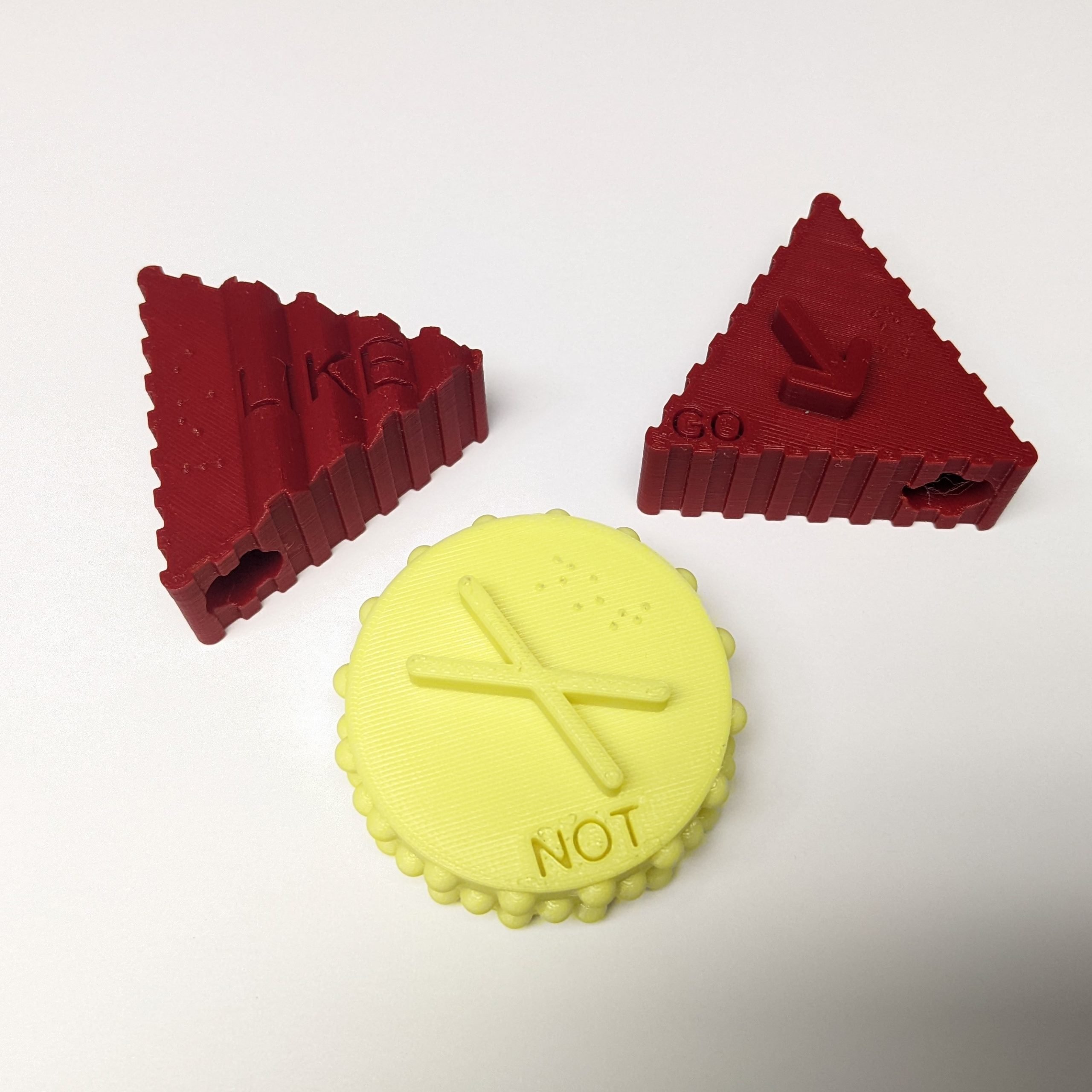 Two red and one yellow 3d printed tactile symbols against a white background. There are three symbols, a triangular red 'Go', a triangular red 'Like', and a circular yellow 'Not'.