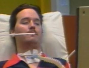 Neil Squire laying in a hospital bed with a tracheotomy tube in his neck and a straw near his mouth