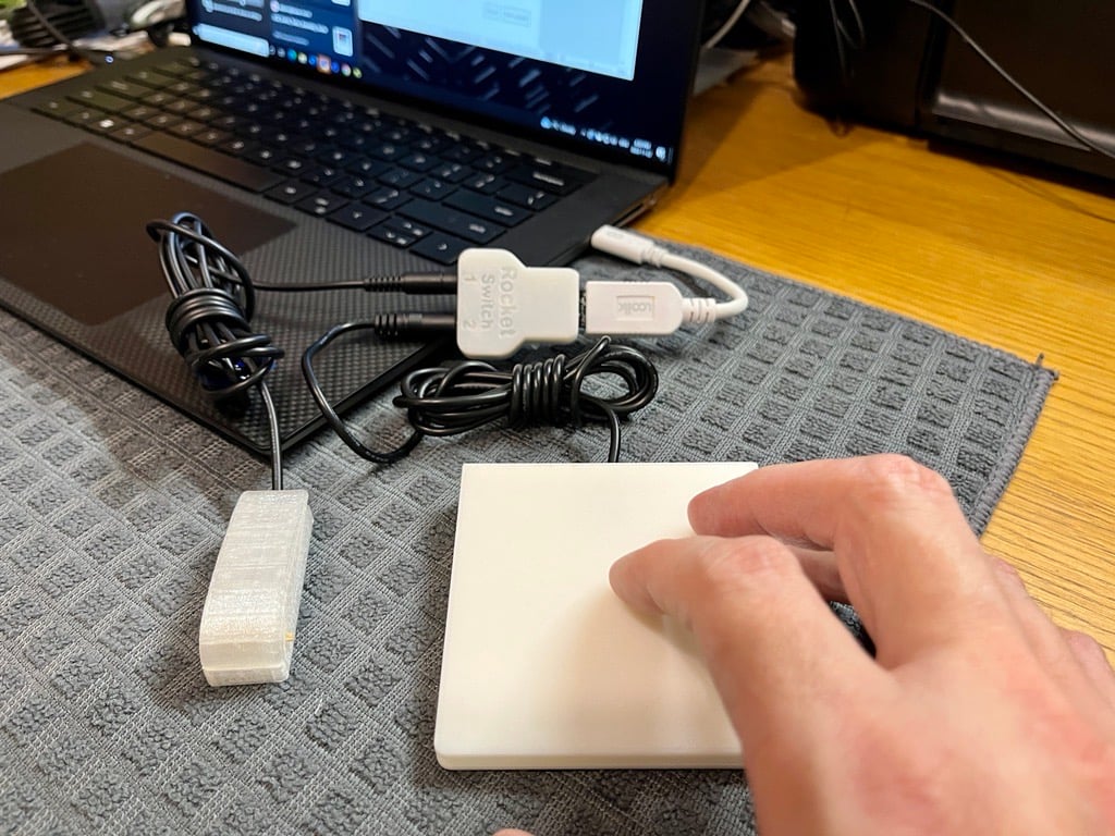 A hand pressing on a white assistive switch plugged into the Rocket Switch, which is plugged into a black laptop.