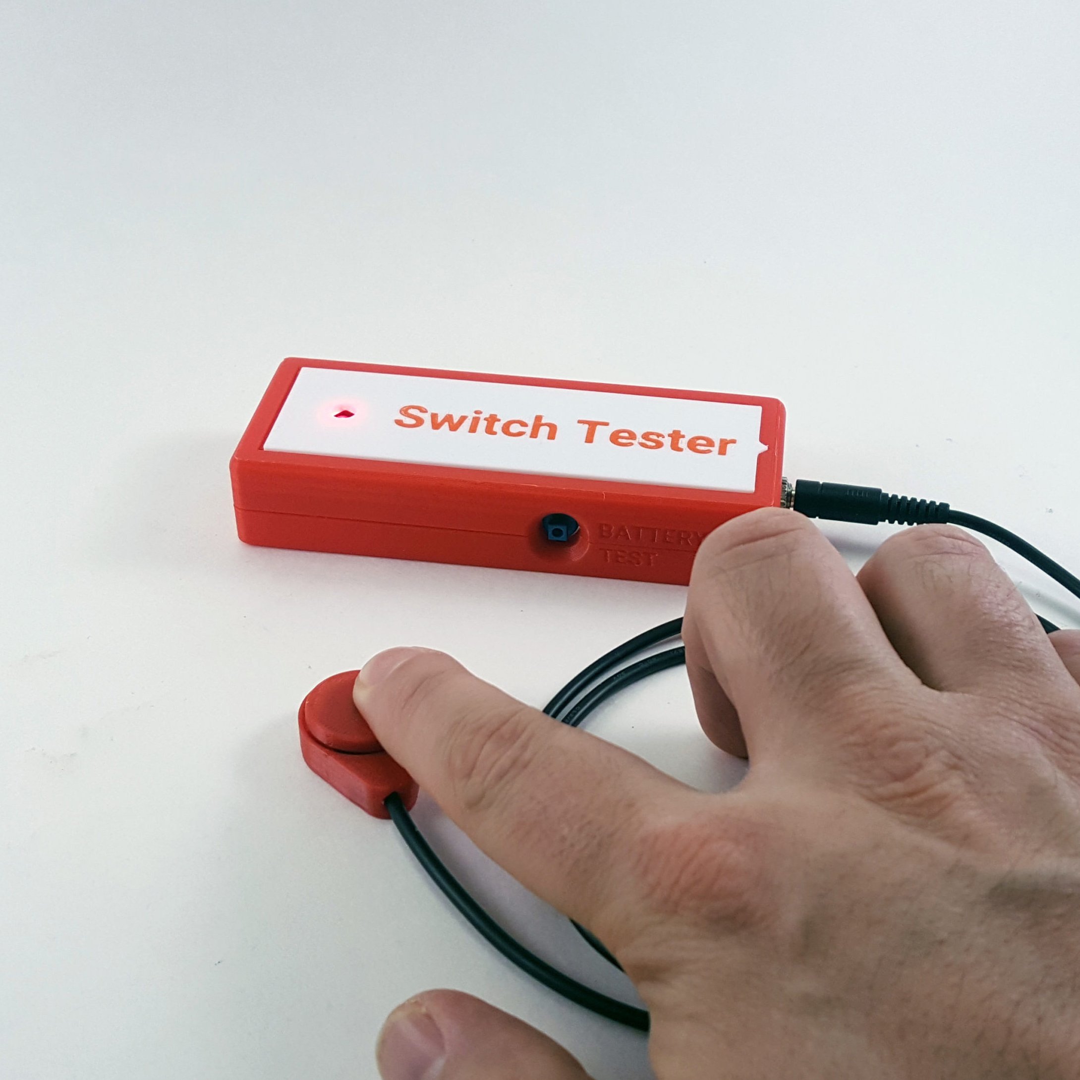 Picture of a 3d printed simple switch tester. A hand is pressing a raindrop switch connected to the tester, which is turning on a red indicator light.