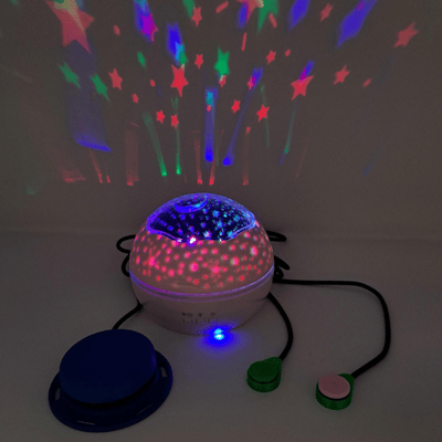 A switch adapted light projector in a dark room, showing colourful stars projected onto the wall beside it. There are 3 different assistive switches plugged into it.