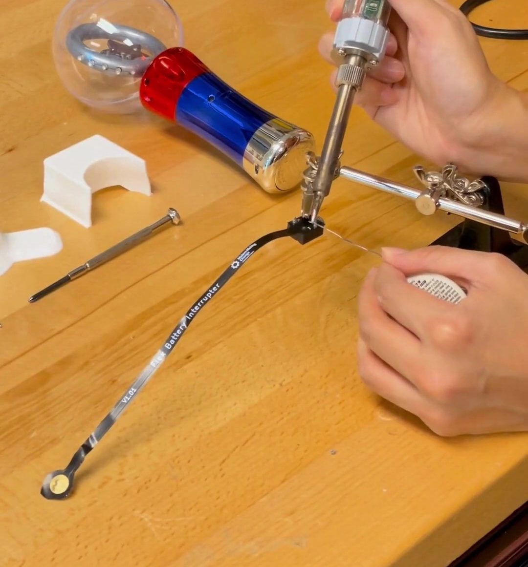 A pair of hands soldering the battery interrupter on a wooden bench.