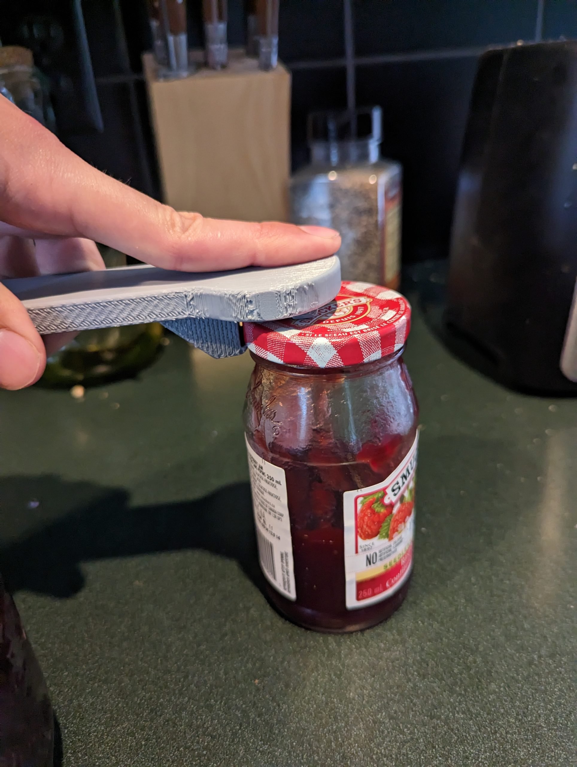 A grey 3d printed jar lid opener hooked on to jam jar lid to break the seal.