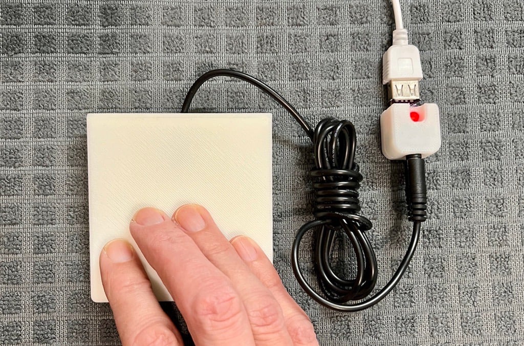 A hand pressing on a white 3d printd assistive switch that's plugged into the USB switch tester, with the red LED on the tester lit up.