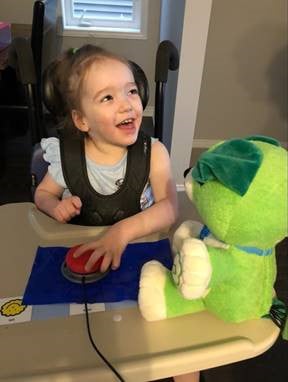 A toddler girl in a high chair laughing and playing with an adapted toy dog