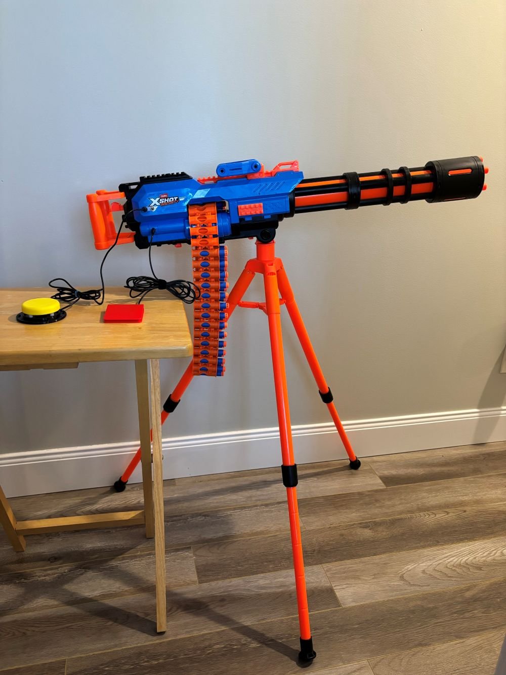 A large blue and orange dart blaster on an orange tripod that has been switch adapted. Two assistive switches are plugged into the dart blaster, one is red and one is yellow.