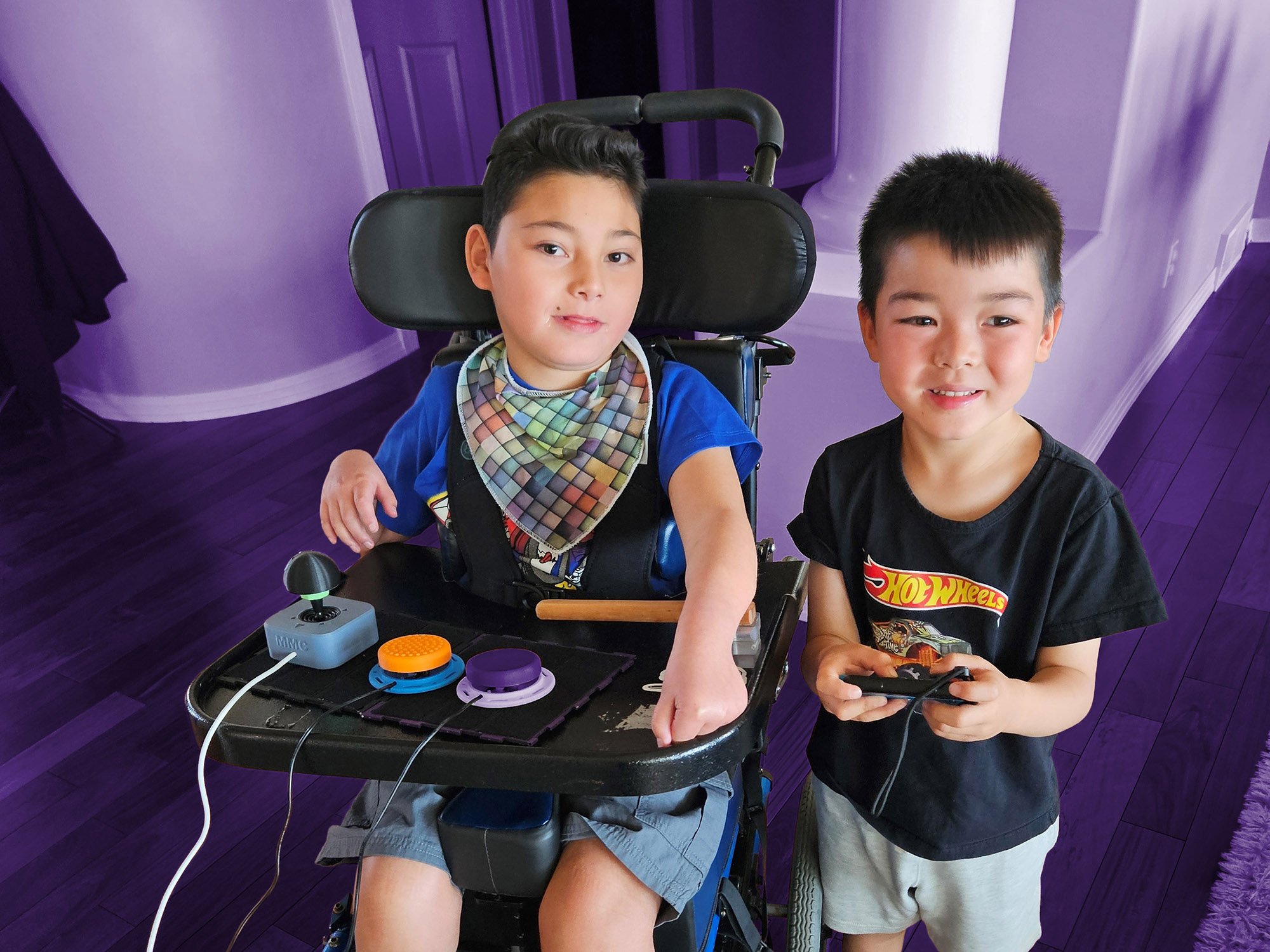 Two boys side by side at home. The boy on the left is in a wheelchair with 3d printed joystick and switches in front of him. The boy on the right is holding a gaming controller