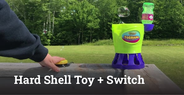 A hand pressing on an assistive switch to activate a bubble blower. Overlaid is the text "hard shell toy + plush" in white.