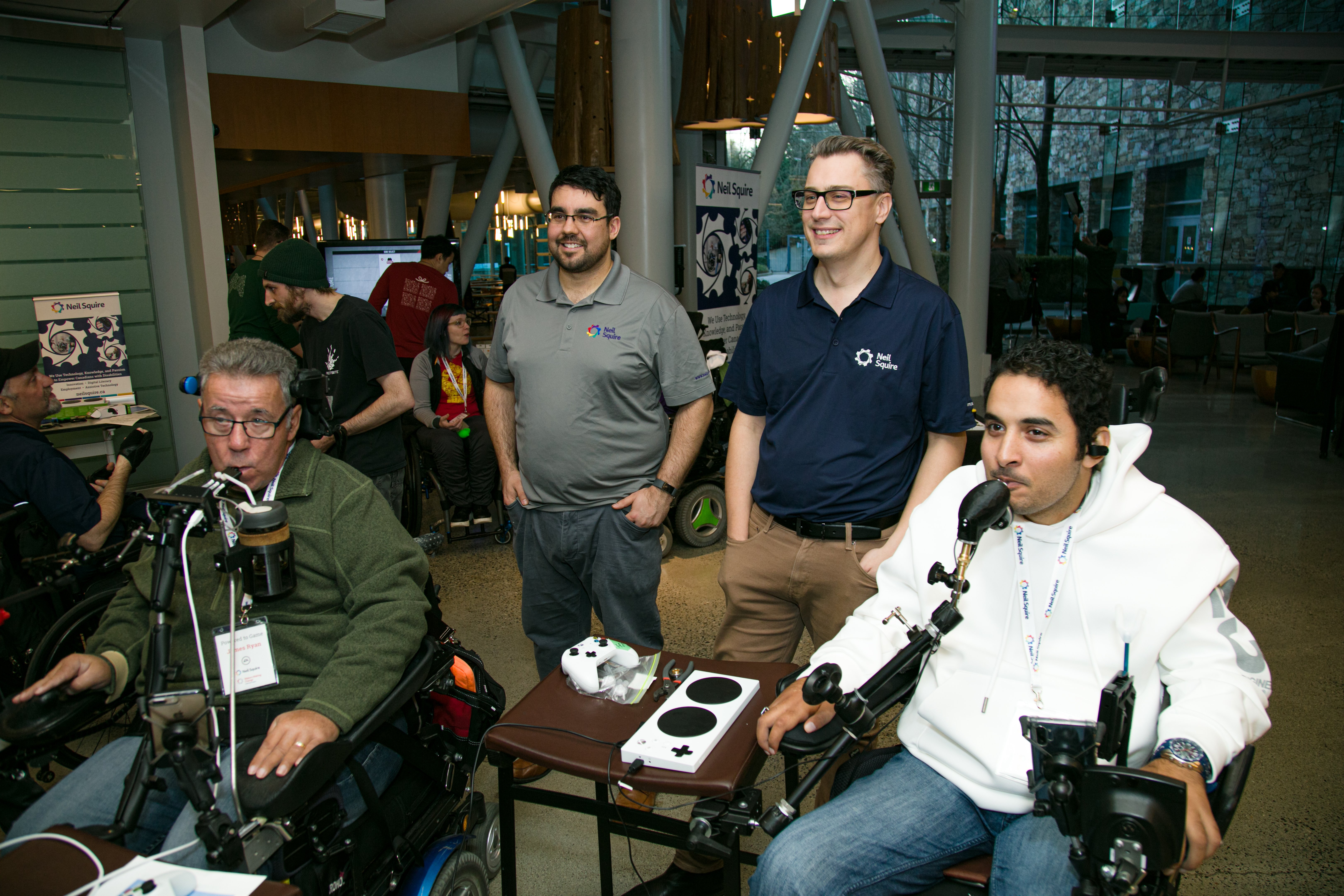 2 gamers in wheelchairs playing video games off screen. Behind them stands 2 makers making change staff