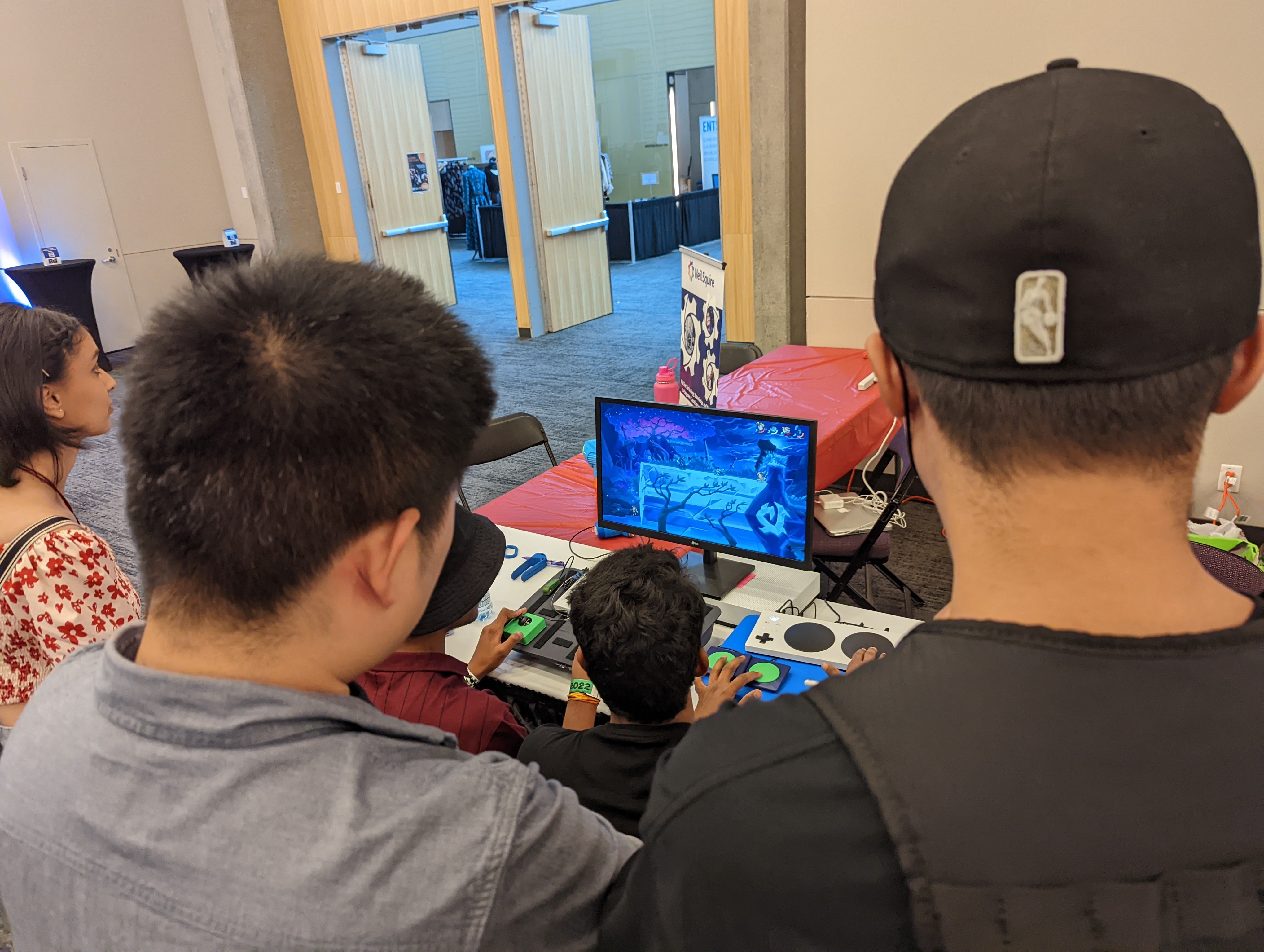 The back of the heads of a crowd of 3 people watching two people play a video game using assistive technology at a booth during an event.