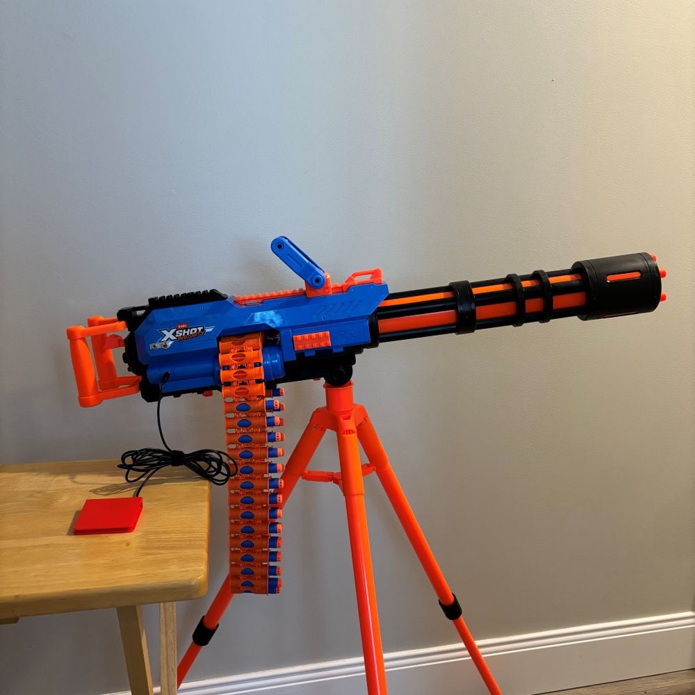 A large blue and orange dart blaster on an orange tripod that has been switch adapted. A red assistive switch is plugged into the dart blaster.
