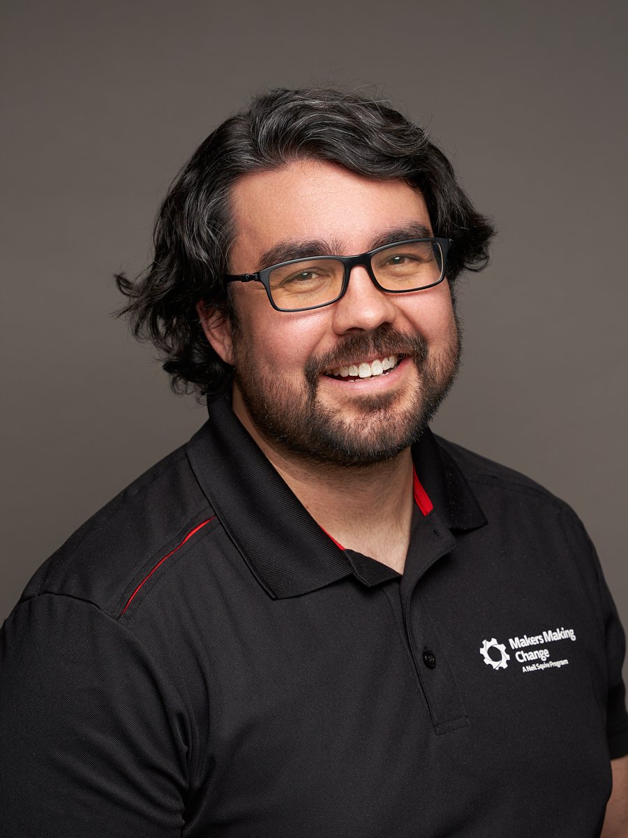 A man with black wavy hair and a black Makers Making Change polo shirt against a grey background.