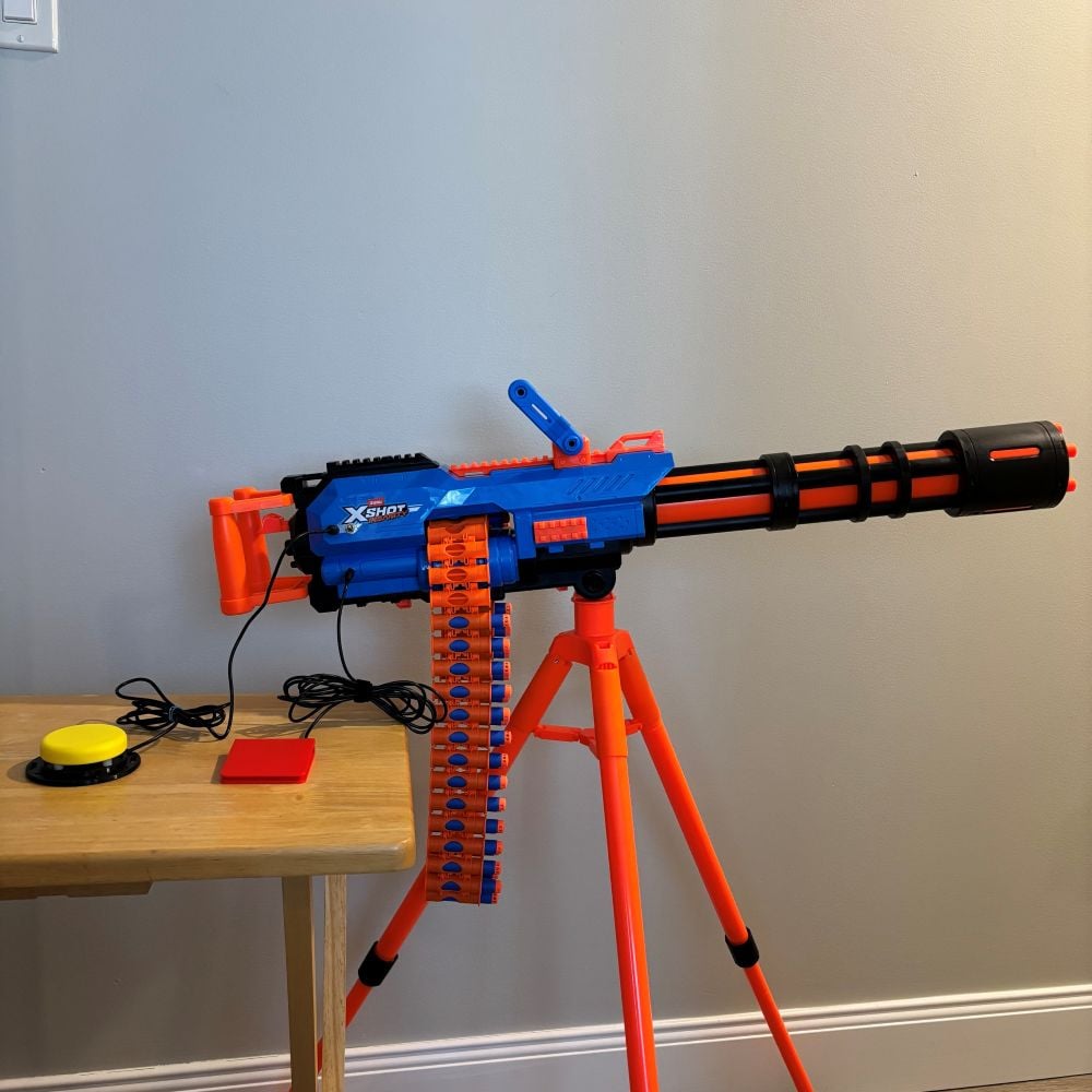 A large blue and orange dart blaster on an orange tripod that has been switch adapted. Two assistive switches are plugged into the dart blaster, one is red and one is yellow.