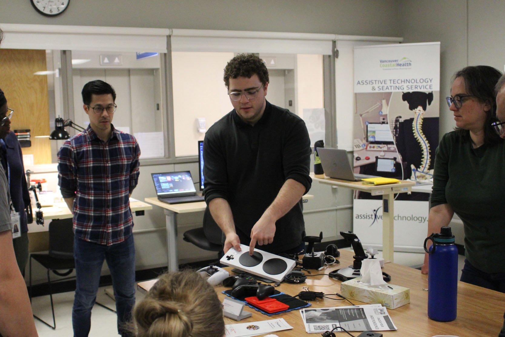 A group of people around an MMC staff member who is pointing at an Xbox Adaptive Controller and explaining how it works.