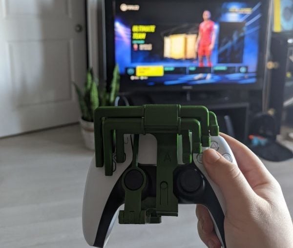 Holding a PS5 controller with a 3d printed controller mod attached. A FIFA video game is on the TV in the living room background.