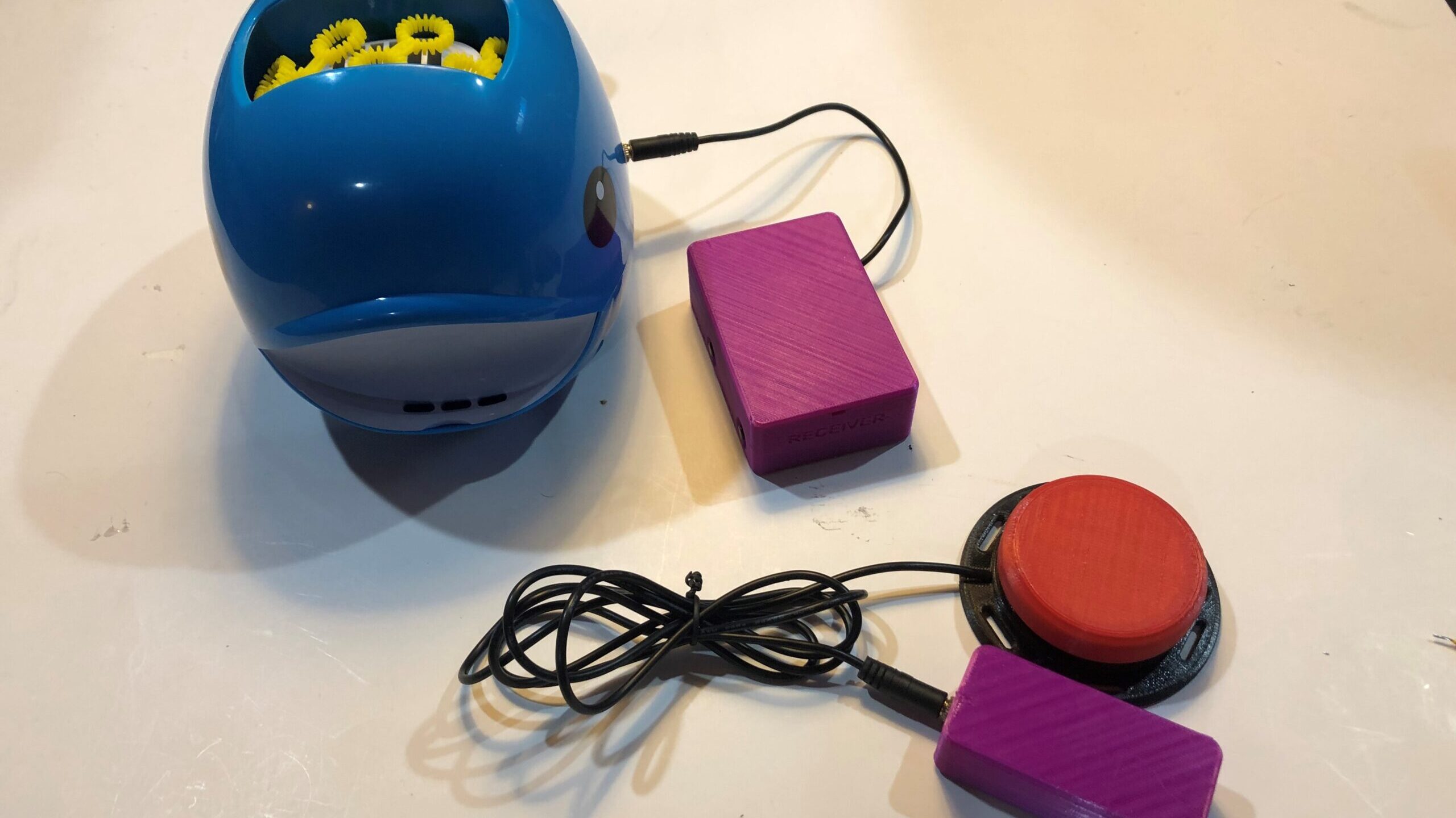 A purple wireless assistive switch link plugged into a red assistive switch and a blue whale bubble blower toy.