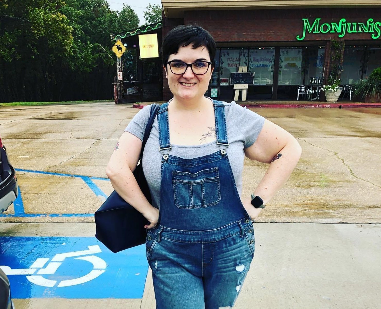 A women wearing a grey t-shirt and denim overalls posing with her hand on her hips.