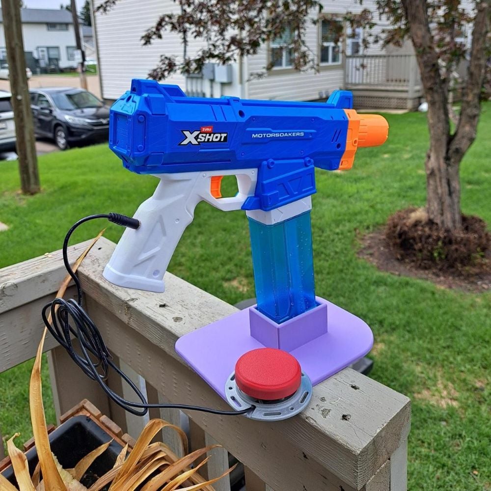 A switch adapted water gun standing up in a purple 3D printed stand. A large red assistive switch is plugged into it in the handle. The water gun is blue and white with an orange nozzle.