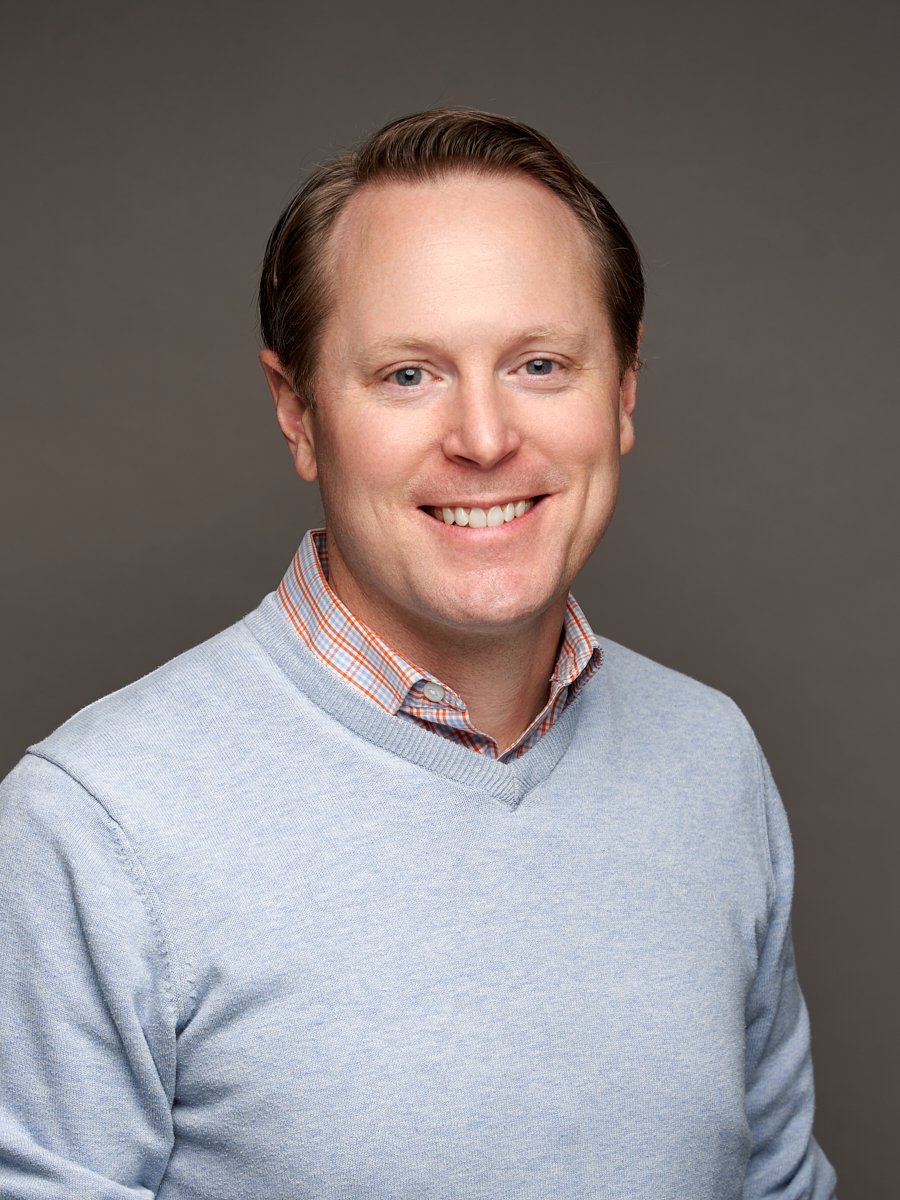 A man with brown hair and light blue sweater against a grey background.
