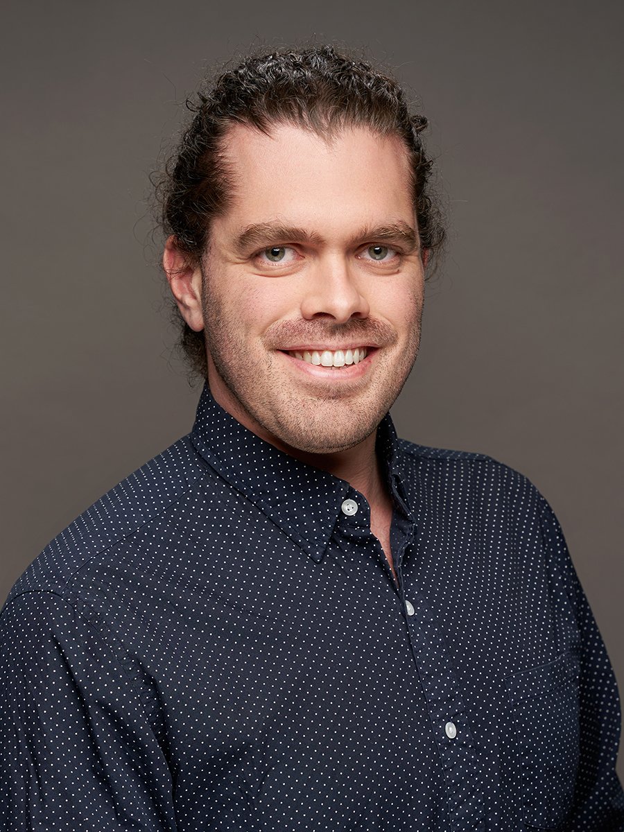 A man with brown tied up hair and blue button up shirt against a grey background.