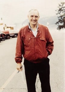 A older man wearing a red jacket standing outdoors, holding a pen in his hand