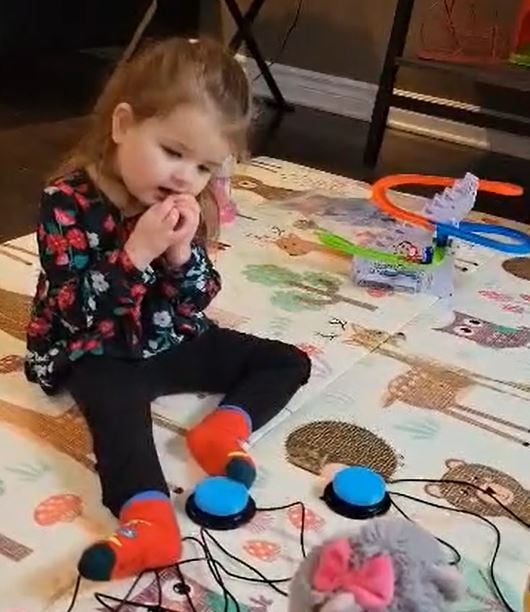 A young girl sitting on the floor using her feet to activate an assisistive switch.