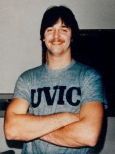 A man wearing a grey t-shirt that says UBC smiling with his arms crossed
