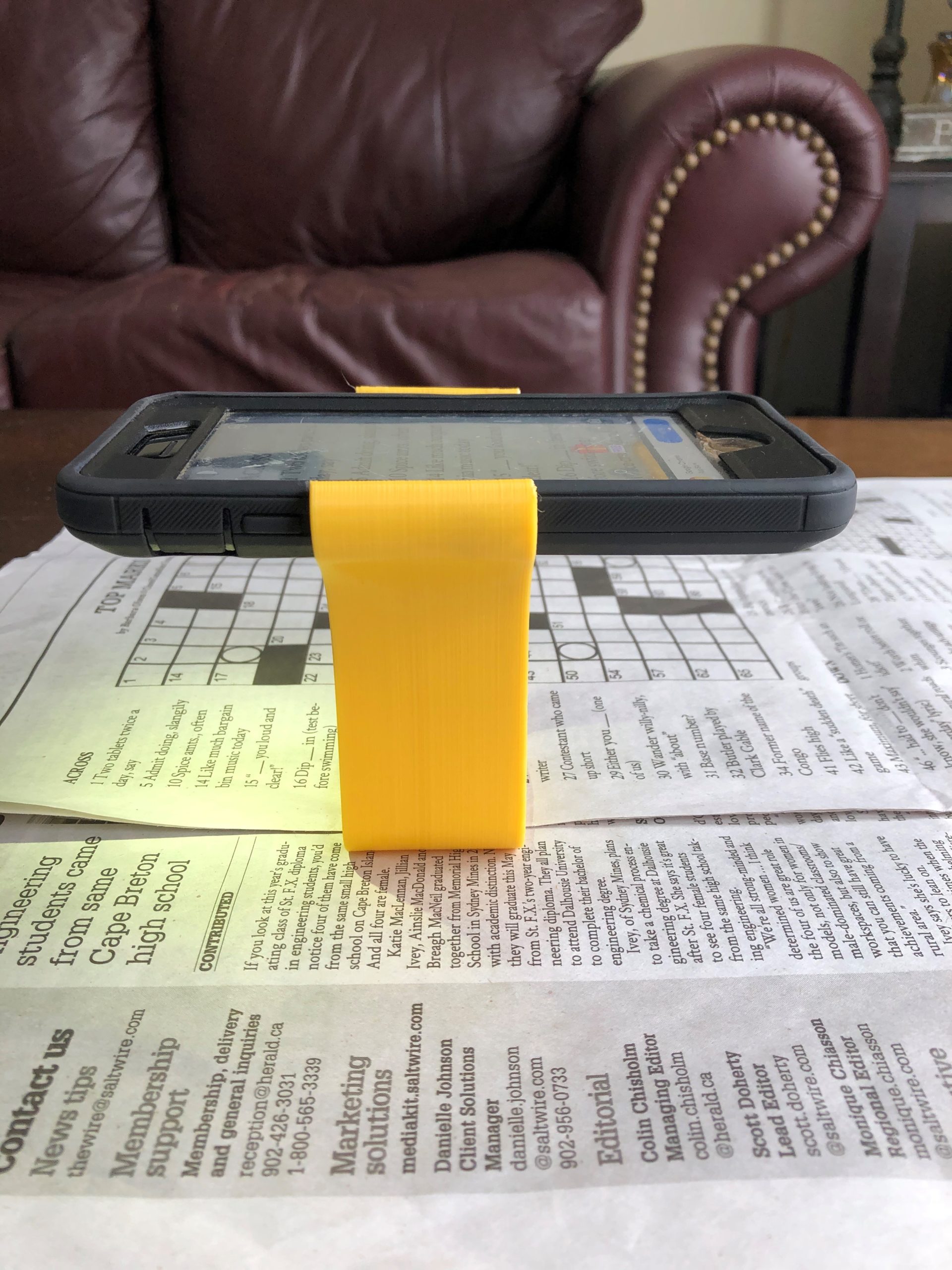 A yellow 3d printed magnification stand holding a phone. The camera app is displaying a zoom in view of a newspaper underneath.
