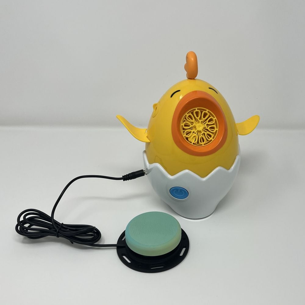 A switch adapted bubble blower that looks like a yellow chick in half an egg. The chick has its beak open for bubbles to come out. There is a large green assistive switch plugged into the toy.