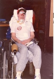 Photo of a man in a wheelchair with sunglasses and a red hat, there are bandages and tubes around his throat