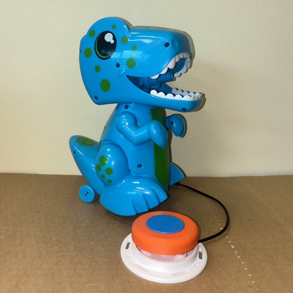 A blue dinosaur toy with an orange and blue assistive switch plugged into it. The dinosaur is cartoon looking and has a spout in its mouth where bubbles come out.