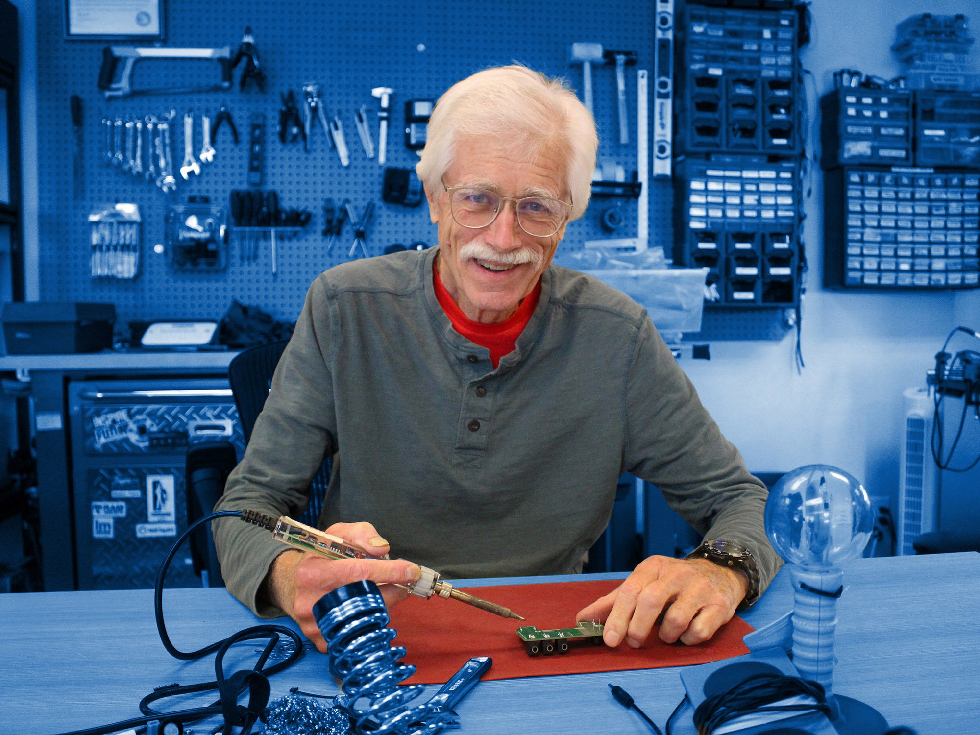 A man soldering electronics with a wall of tools behind him