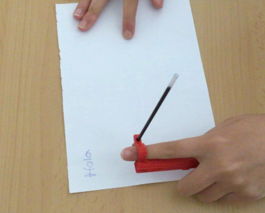 A person using a red 3d printed arm-mounted pen holder to write "Hola" on a piece of paper.